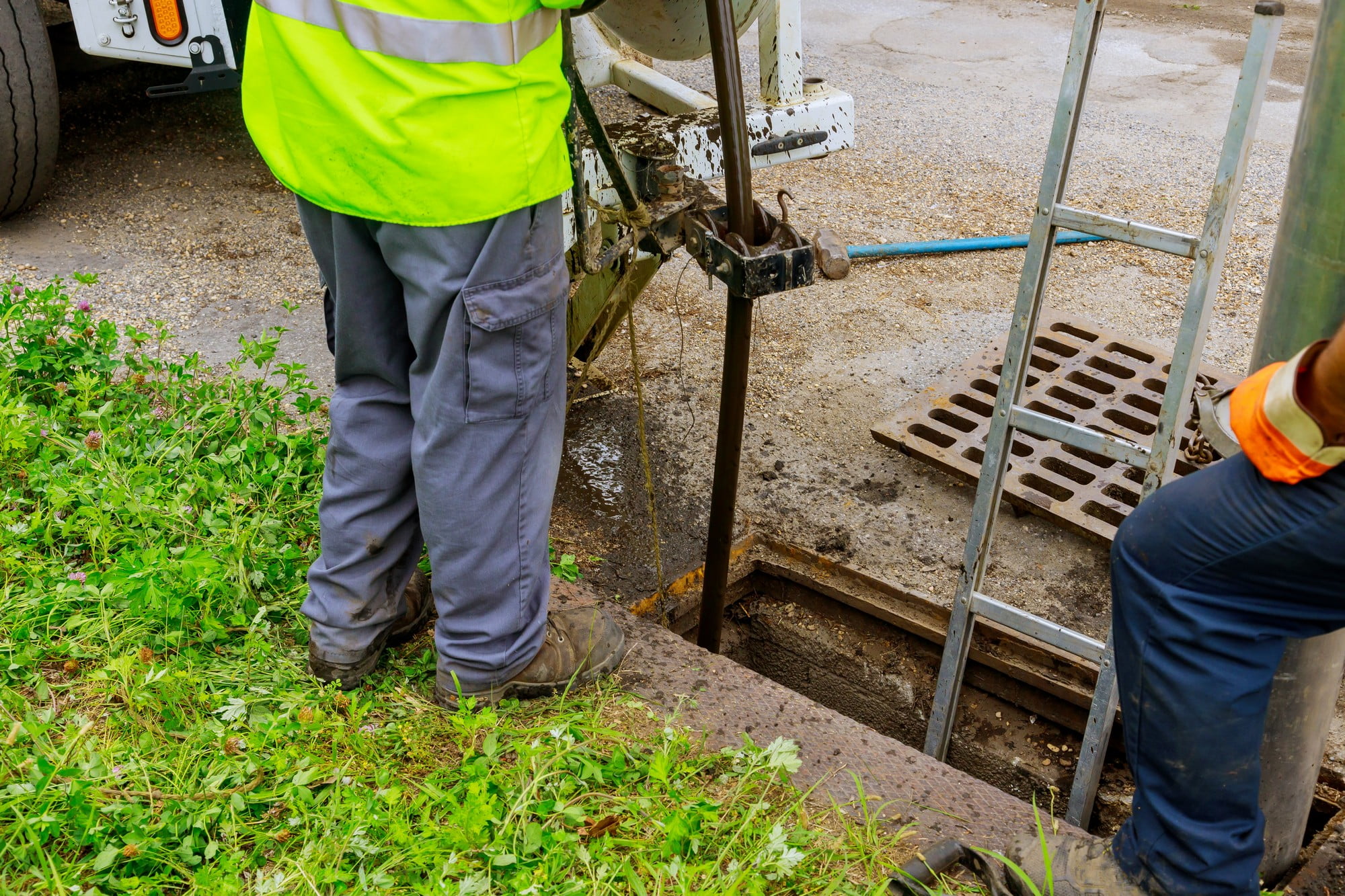 In the image, a worker is engaged in cleaning the sewer system using specialised equipment. The scene is set outdoors near a utility access point. The worker, dressed in bright orange high-visibility protective clothing, including a hard hat and gloves, is operating a large, lorry-mounted machine designed for sewer cleaning. The machine has long, flexible hoses that are being inserted into a manhole, through which the cleaning process occurs. Nearby, orange traffic cones are placed around the work zone to ensure safety and highlight the area to pedestrians and traffic. The background shows an urban environment with buildings and greenery, indicating that the sewer maintenance is taking place in a public or residential area. The overall setting emphasizes the importance of safety and efficiency in public utility maintenance.